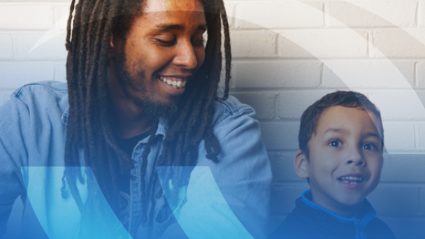 A man with black, shoulder length dreads is wearing a blue oxford button down and is smiling down at a small child who sits next to them. The child has short brown hair and wears an amused and happy expression.