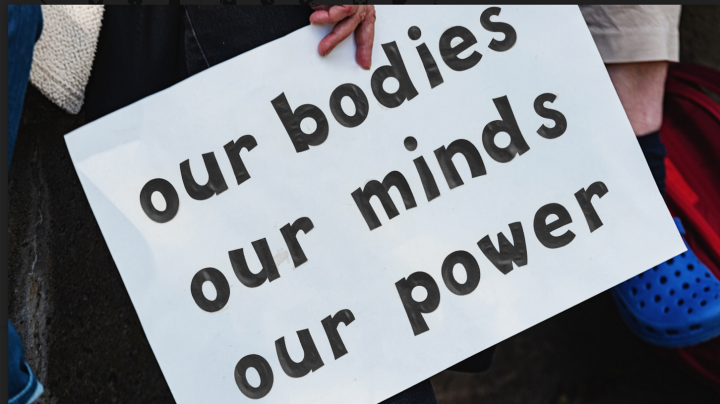 Banner with white background being held by a person's hand. Banner reads "our bodies", "our minds", "our power".
