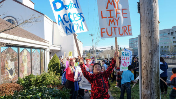 Young kids are protesting for dental access. They hold homemade signs that say "Dental Access Now" and "My Life My Future" - Photo credit to Children's Alliance in Seattle, WA