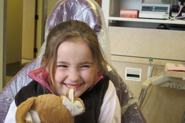 A young child is grinning while sitting in a dentist's chair and squeezing a stuffed animal.