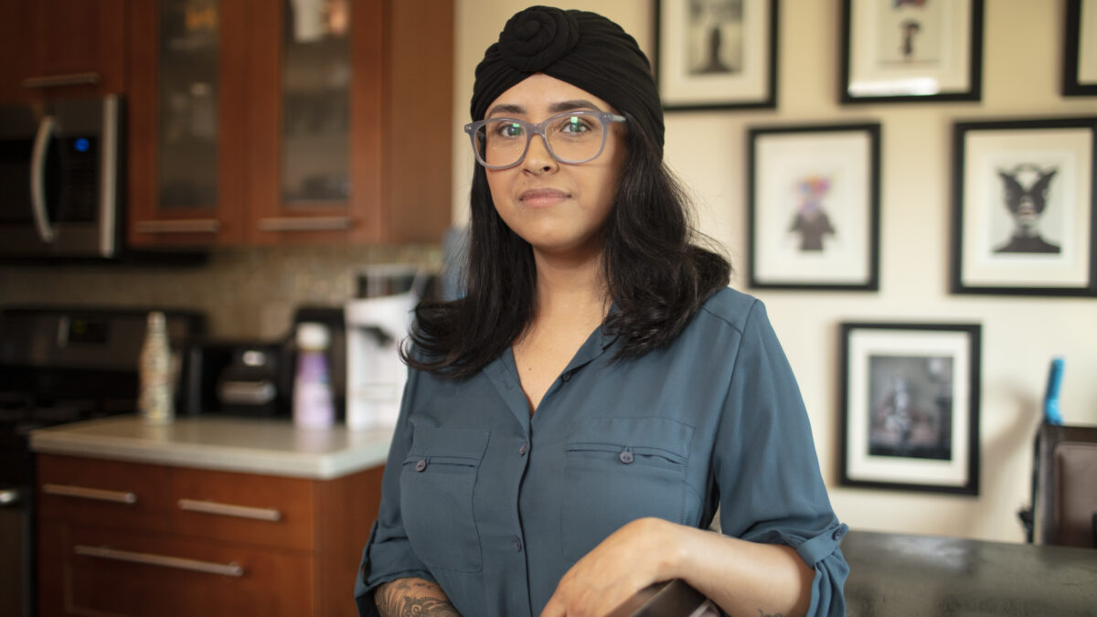 Aracely is standing in their home next to a chair, wearing a dark teal button-down blouse with rolled up sleeves to reveal a delicate tattoo that covers most of their right forearm. Kitchen cabinets and a countertop, and a collection of art in black frames on a wall, are in the background. Aracely, who wears large, light gray glasses, has dark, shoulder-length hair and wears a black-knotted headscarf that covers part of their head. Aracely has a slight smile with a closed mouth.