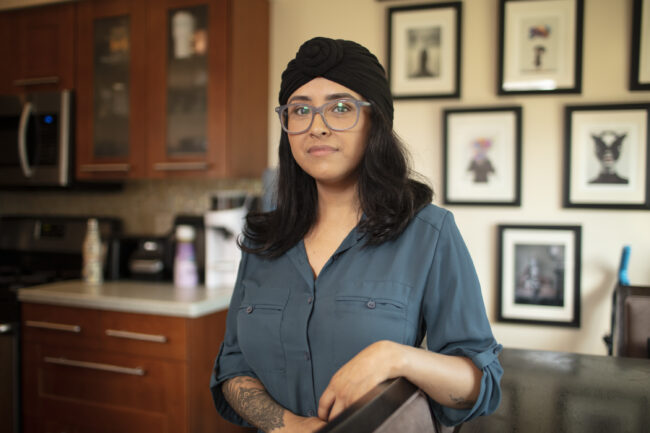 Aracely is standing in their home next to a chair, wearing a dark teal button-down blouse with rolled up sleeves to reveal a delicate tattoo that covers most of their right forearm. Kitchen cabinets and a countertop, and a collection of art in black frames on a wall, are in the background. Aracely, who wears large, light gray glasses, has dark, shoulder-length hair and wears a black-knotted headscarf that covers part of their head. Aracely has a slight smile with a closed mouth.