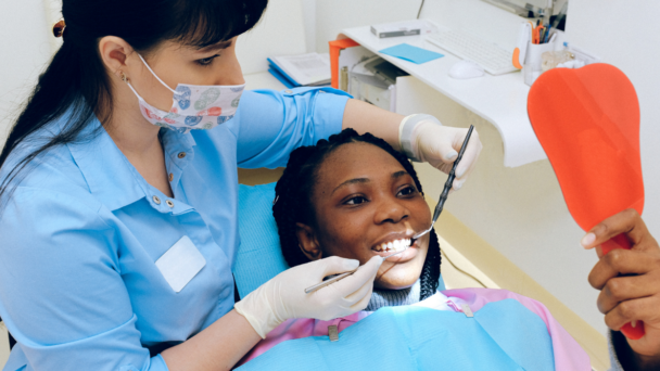 A patient, with hair in twists, lies back in a dental chair while holding a red hand mirror. The patient is smiling as the oral health technician, in blue with a floral face mask, is working on her teeth.