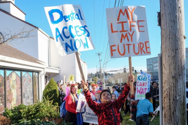 People are gathered at a protest with signs that say "Dental Access Now" and "My Life My Future"