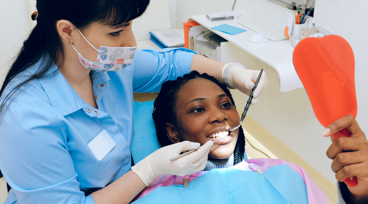 A patient, with hair in twists, lies back in a dental chair while holding a red hand mirror. The patient is smiling as the oral health technician, in blue with a floral face mask, is working on her teeth.