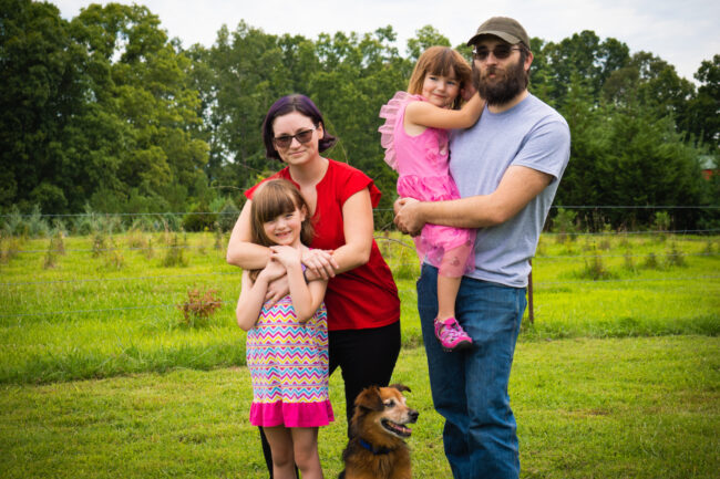 A family of four, plus a brown dog, smiles for a photograph in their green yard in a rural setting. From left to right: young child with red hair and bangs, pink stripes; mother in red shirt and sunglasses and armed wrapped around the child; small child in pink is being held by their father; father, in a light t-shirt and jeans, wears a full beard and a baseball cap.