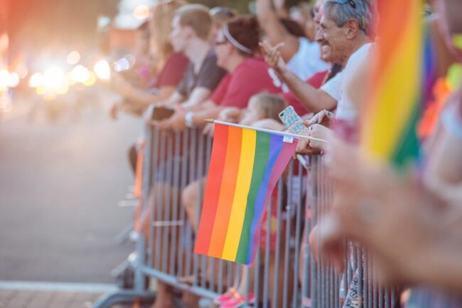 A group of people appear to be behind metal barricades to watch a parade. There is a gay pride rainbow flag, and everyone appears to be in good spirits.