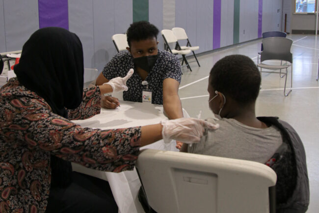 A young child is sitting in a chair with their back to the viewer. They are wearing a face mask and their shirt sleeve is rolled up, as they are about to get a vaccine. Two health workers are assisting; one to their left, in a black head scarf and paisley shirt, wears gloves and is preparing the vaccine shot. The other, wearing blue patterned medical scrubs, is making direct eye contact with the young person, as if to reassure them.