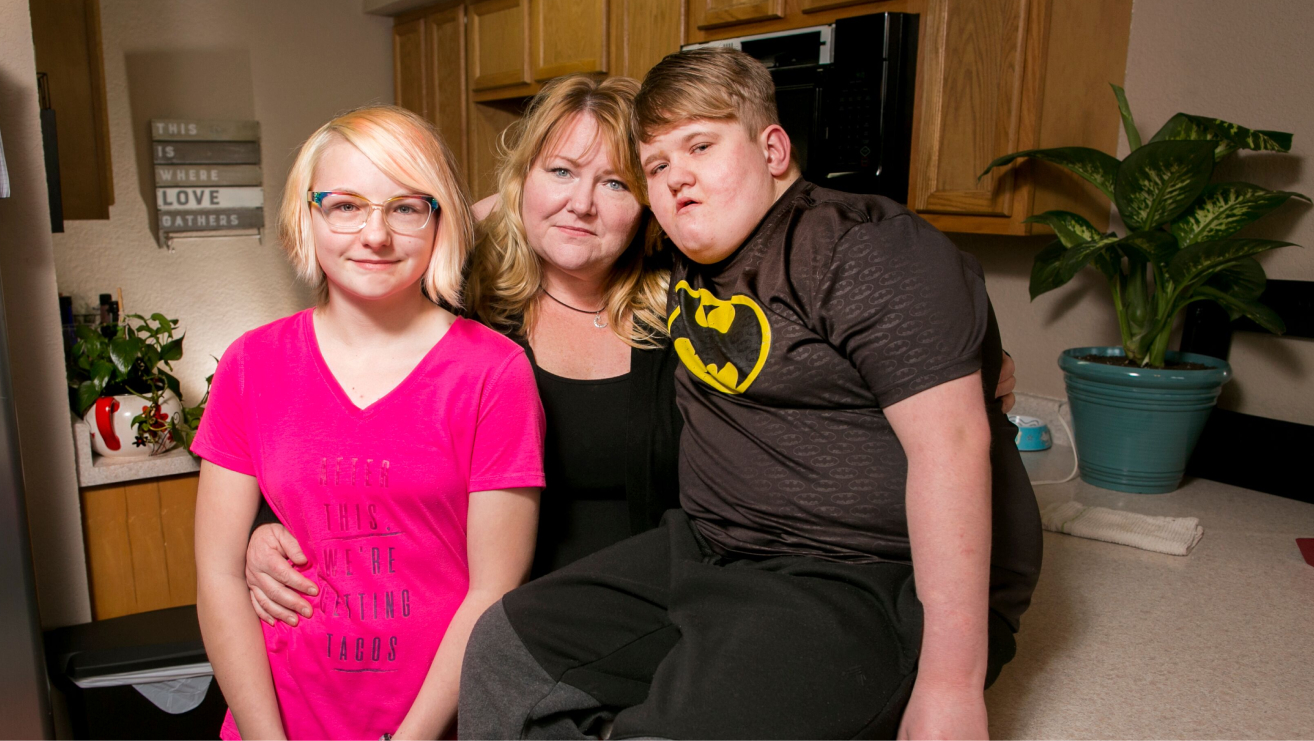 In a kitchen, a mother wraps her arms around two teenage children on either side of her. One teenager has a platinum blonde bob, and wears rainbow-colored glasses and a bright pink t-shirt that says "After this, we're eating tacos.” The mother has sandy blonde hair, styled with bangs and waves, wears a black top, and looks directly at the viewer with light-colored eyes. The teen on the right sits on top of the kitchen counter, has short dark blonde hair, and wears a t-shirt with a Batman logo. A wooden piece of art on the wall is visible and reads: "This is where love gathers.”