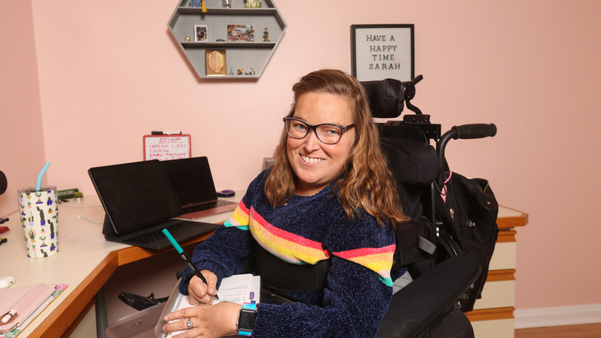 Sarah, a young woman with light brown hair, is wearing black glasses and a wide smile. Sitting in her wheelchair at her desk at work, Sarah has a notepad and pen in hand. Behind here is a small framed sign that says: "Have a happy time Sarah”