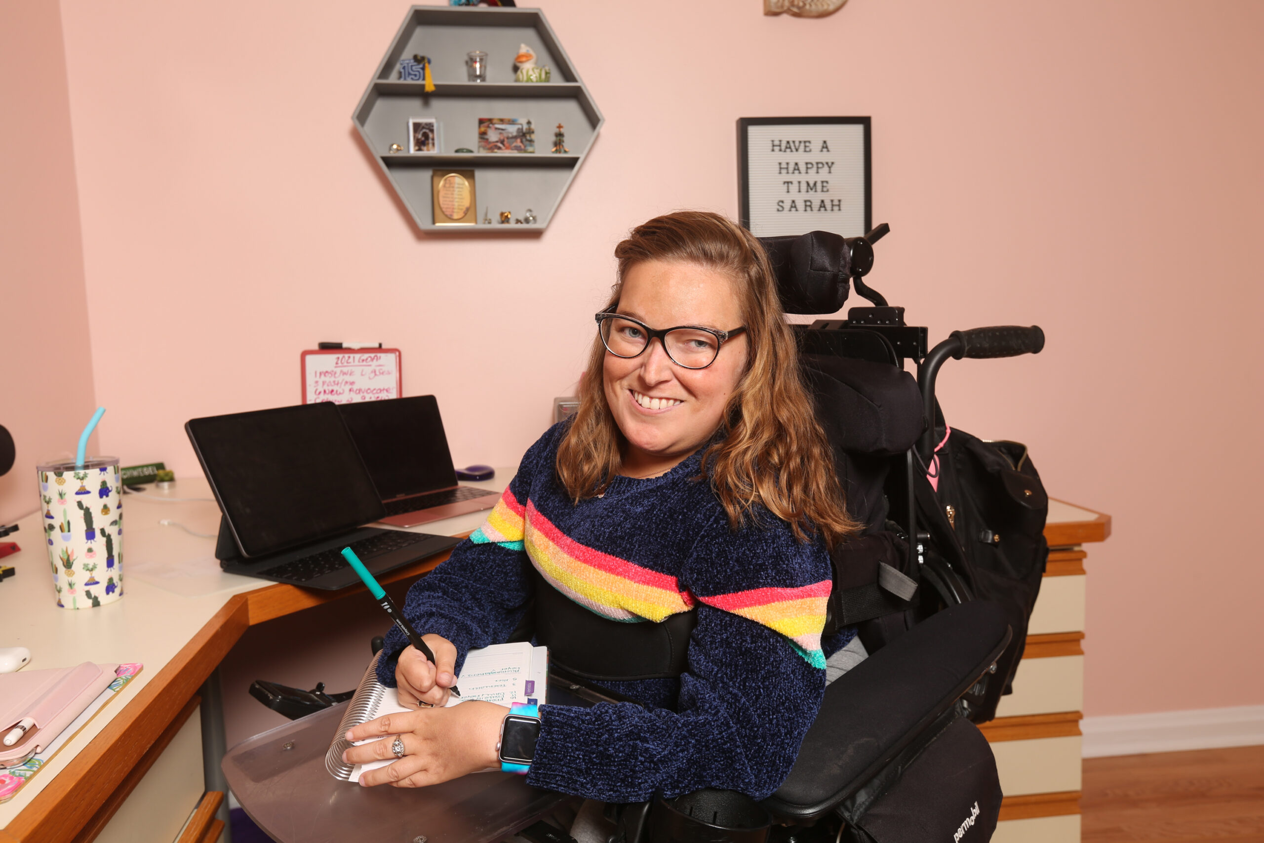 Sarah, pictured, is wearing black glasses and a wide smile. Sitting in her wheelchair at her desk at work, Sarah has a notepad and pen in hand. Behind here is a small framed sign that says: "Have a happy time Sarah"