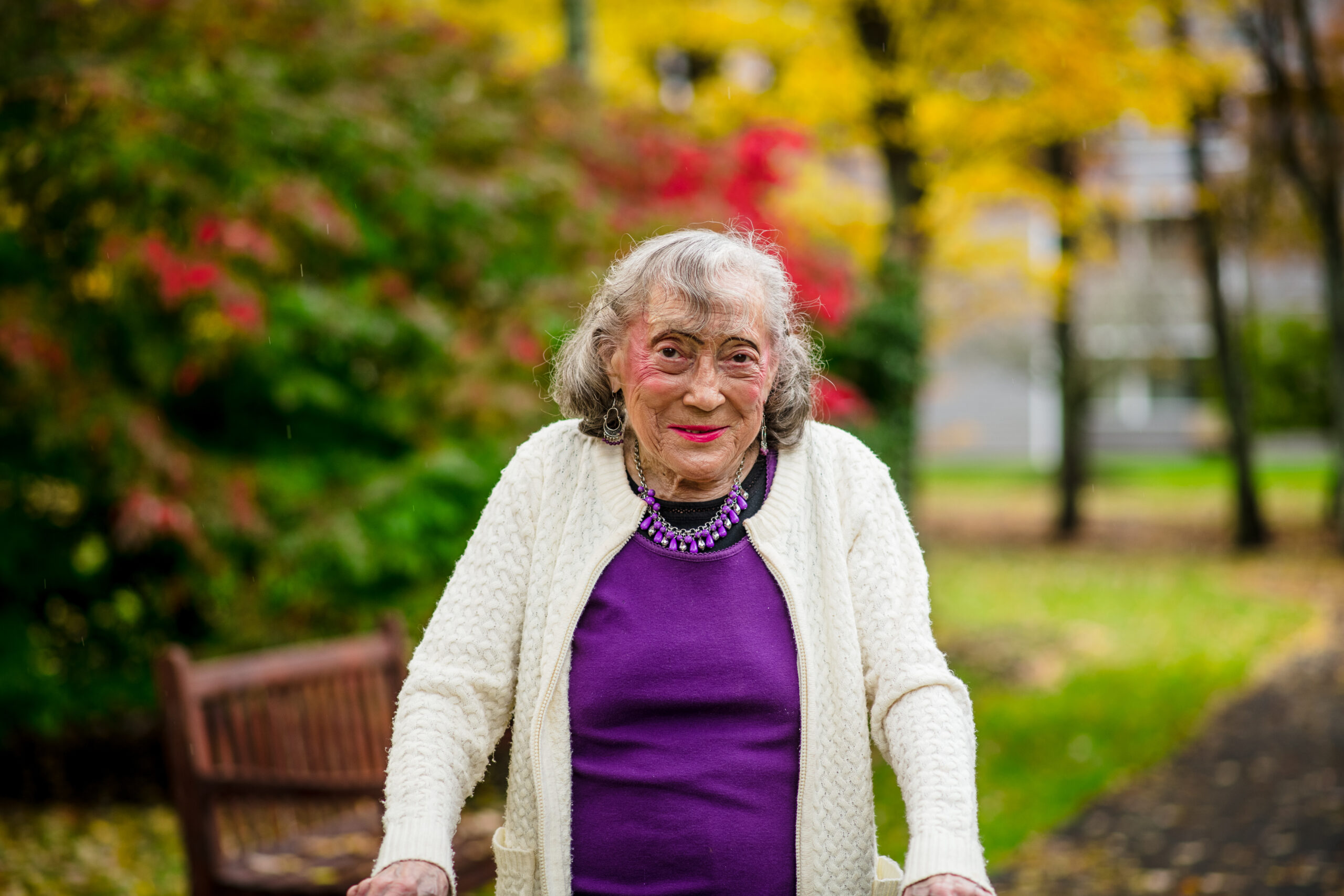Shirly, an older person with bobbed gray hair and wearing a purple shirt and necklace with a cream-colored cardigan, is pictured in a park. Shirly is enjoying the changing colors of the fall leaves, which are brilliant shades of green, red, orange, and yellow. There is a wooden park bench nearby, and green grass and a walking trail are in the background. Whether or not someone has access to neighborhood green spaces, like a local park, is one example of a social determinant of health. 
