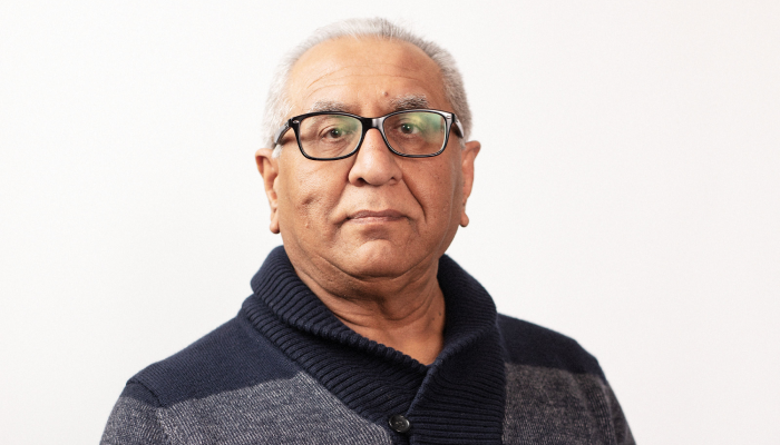 Mahesh Bhatia wearing a navy blue and grey shawl cardigan against a light background looks directly at the camera.