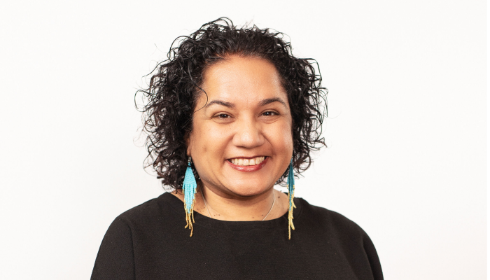 Stacey Wright smiles directly at the camera in front of a light background wearing a black top and turquoise earrings.
