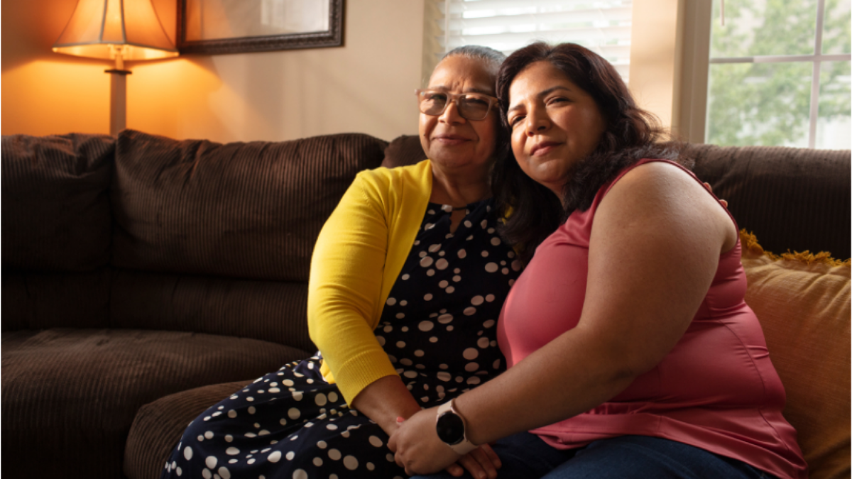 Suzy sits on the sofa with her mother, Angelina, in an embrace. Angelina wears a yellow cardigan over a patterned long dress. Suzy wears a salmon pink sleeveless blouse. The room in warmly lit. The family overcame medical debt thanks to charity care.