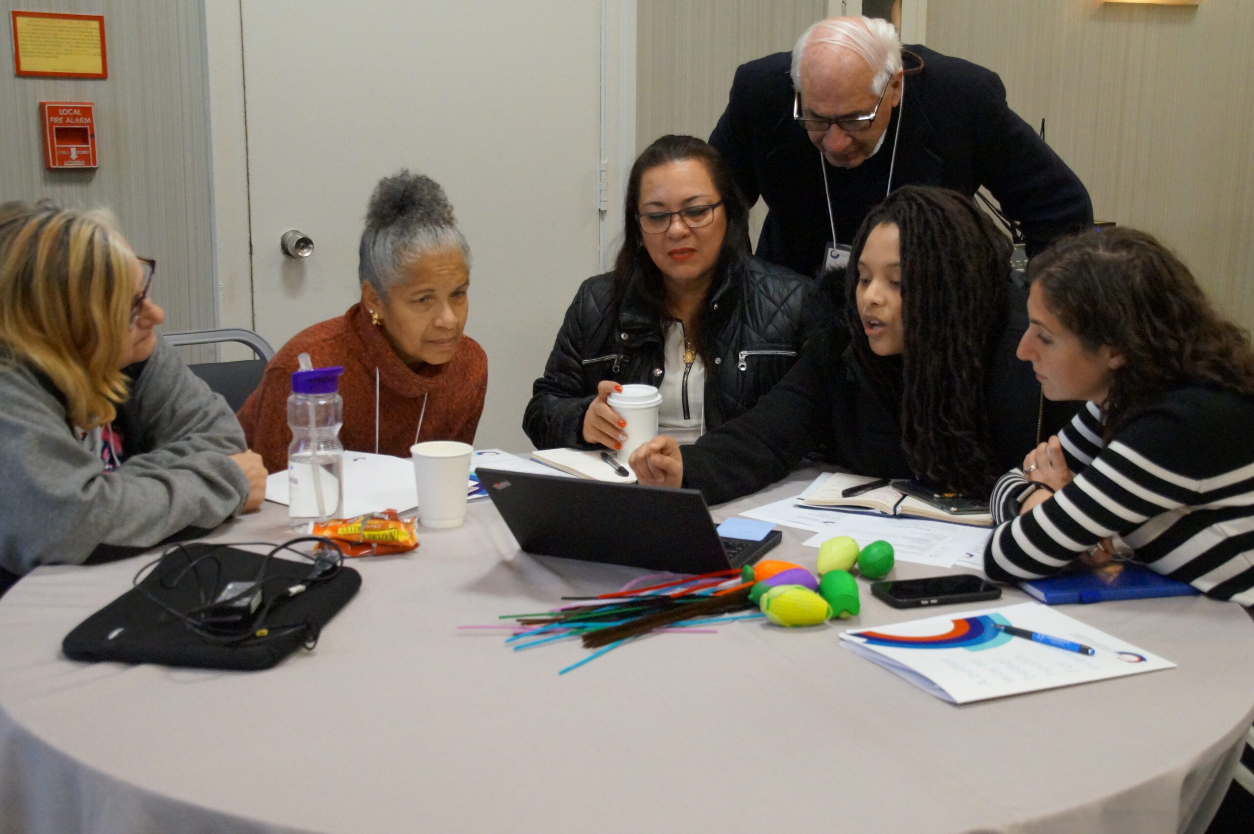 Six health advocates gather around a table to collaborate. They are all looking at a shared laptop screen while one person points to the screen itself, as if sharing thoughts with the group.