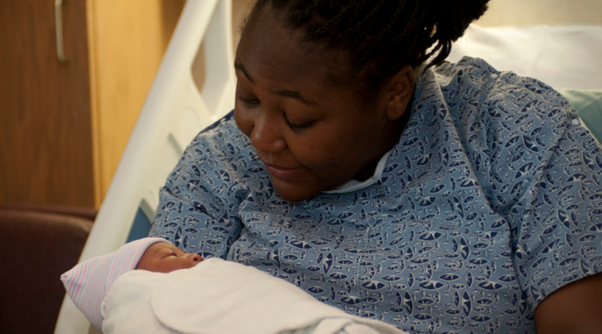 A woman in a blue hospital gown is gazing intently at her newborn child, in her arms, while in a hospital bed.