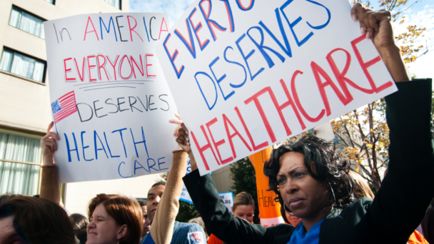 Two activists holding posters above their heads that say “In America Everyone Deserves Healthcare”