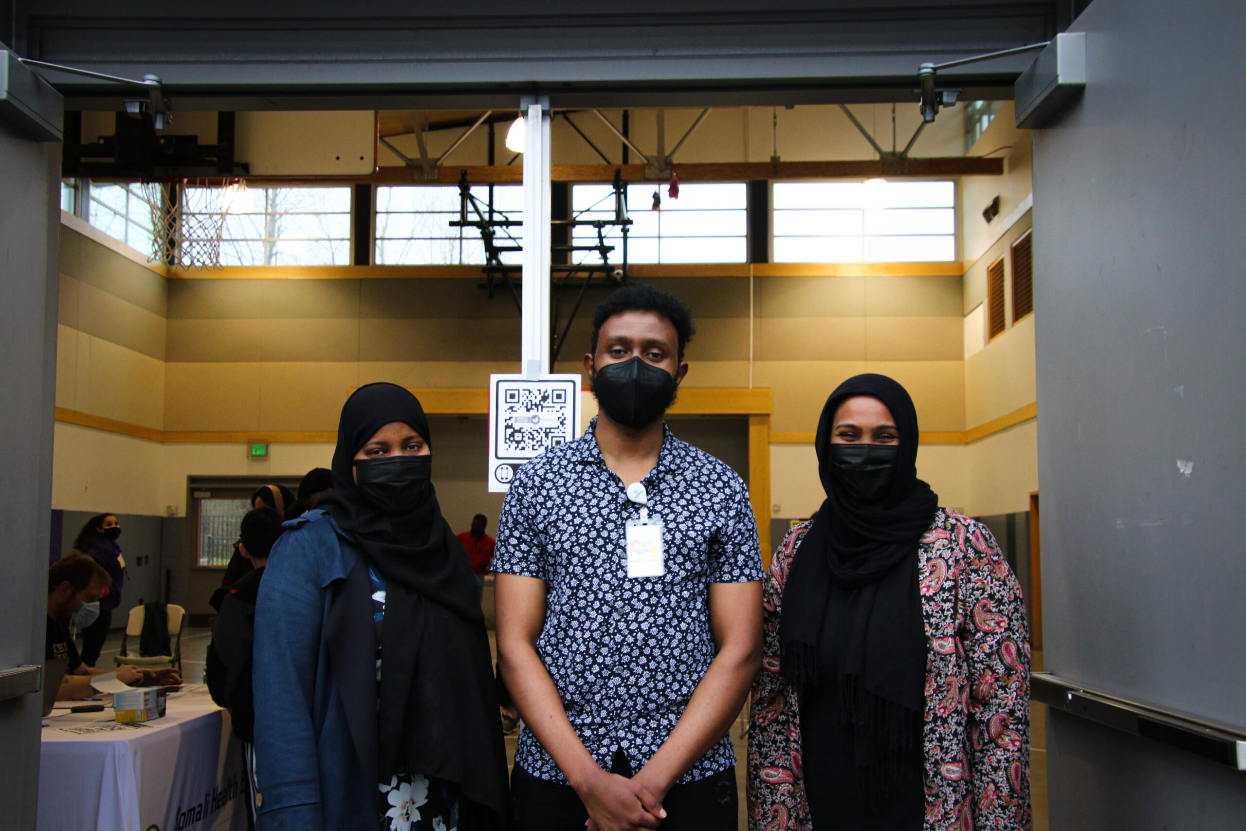 Abdifatah Ali Dahiye, in a patterned short sleeve button-up and a mask, stands in between two colleagues who are wearing masks and head scarves. They are at a public event for COVID-19 testing.