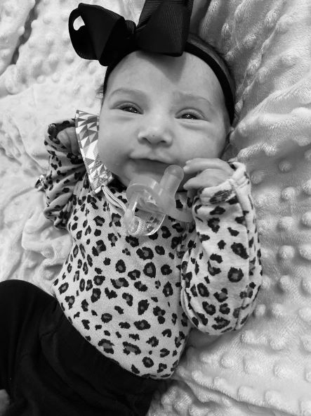 In black and white, a small baby girl in a leopard print onesie and a large black headband with a bow.