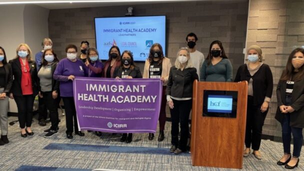 Group of people pose for a picture in a front of a TV display. Three people in the front hold a big sign that reads Immigrant Health Academy. There's a podium to the right.