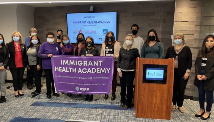Group of people pose for a picture in a front of a TV display. Three people in the front hold a big sign that reads Immigrant Health Academy. There's a podium to the right. 