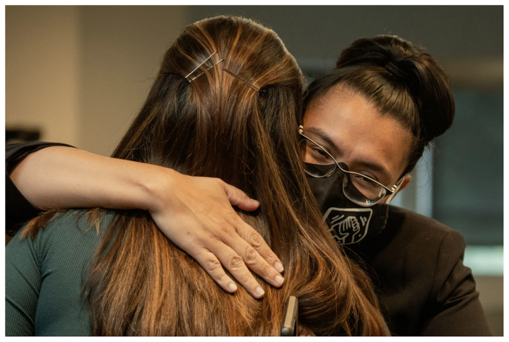 Two people embrace in a hug. One person is facing forward, the other is facing backwards. Person facing backwards has long brown hair. Person facing forward has black hair up in a pony tail, wears a surgical face mask and glasses. Person facing forward has hand over person facing backwards. 