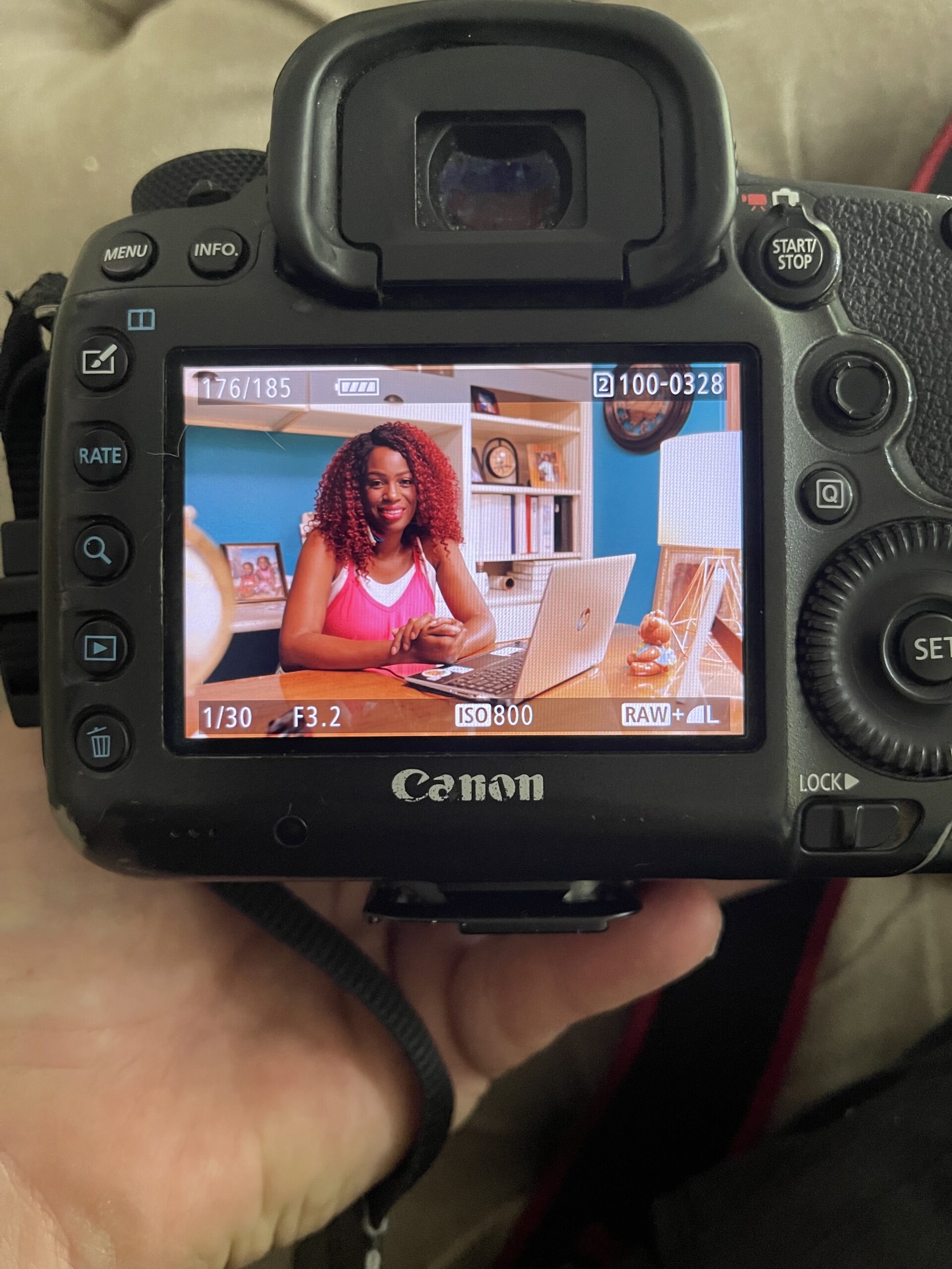 A Canon DSLR camera is held by the photographer, revealing the digital screen and controls. On the screen is a just-captured photo of Sherrell at her desk with a laptop, smiling directly at the camera.