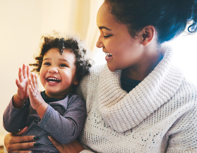 A young mother, in a cream cowl neck sweater, embraces a young child, mid clap with a joyful expression.