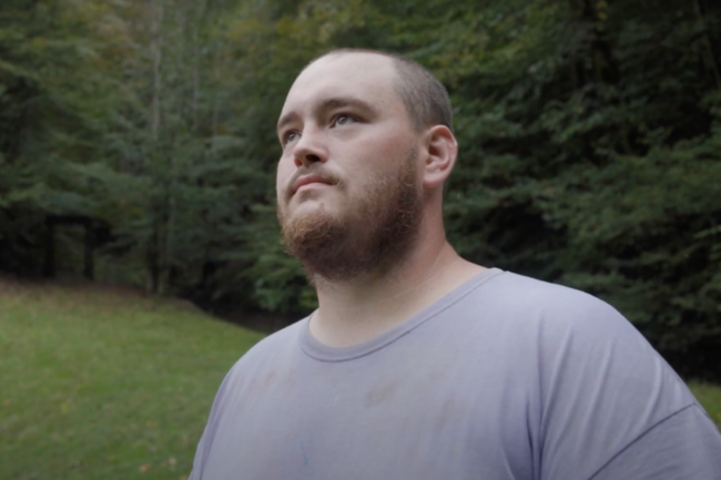 Brandon, who lives and works in Kentucky, is wearing a light gray t-shirt, closely shaved hair, and a full beard.
