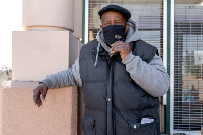 A man wearing a black puffer vest, light grey hoodie sweatshirt, and a cap is wearing a face mask to promote public health and safety during COVID-19.