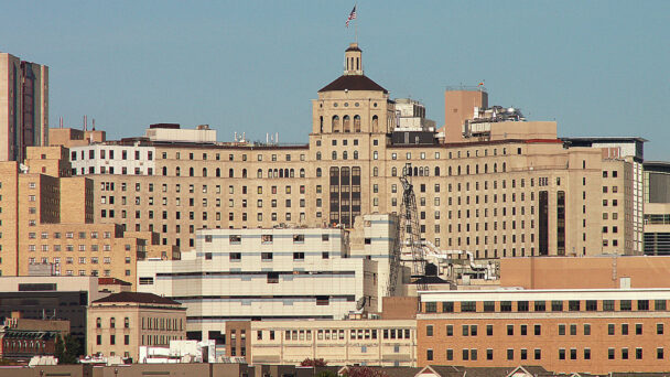 Image of several buildings. At the top sits a big hospital. The hospital building is beige with multiple windows.