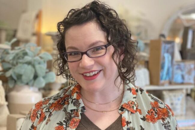 Sarah Farren has brown curly hair, wears black framed glasses, and a patterned floral shirt with a brown v-neck.