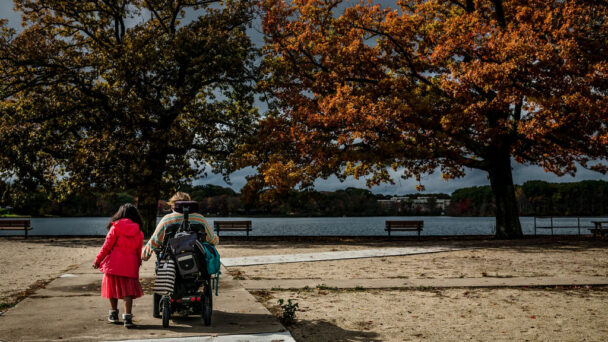 Crystal, a Medicaid-Medicare beneficiary who uses a wheelchair, holds hands with a young girl wearing red in a park by the water