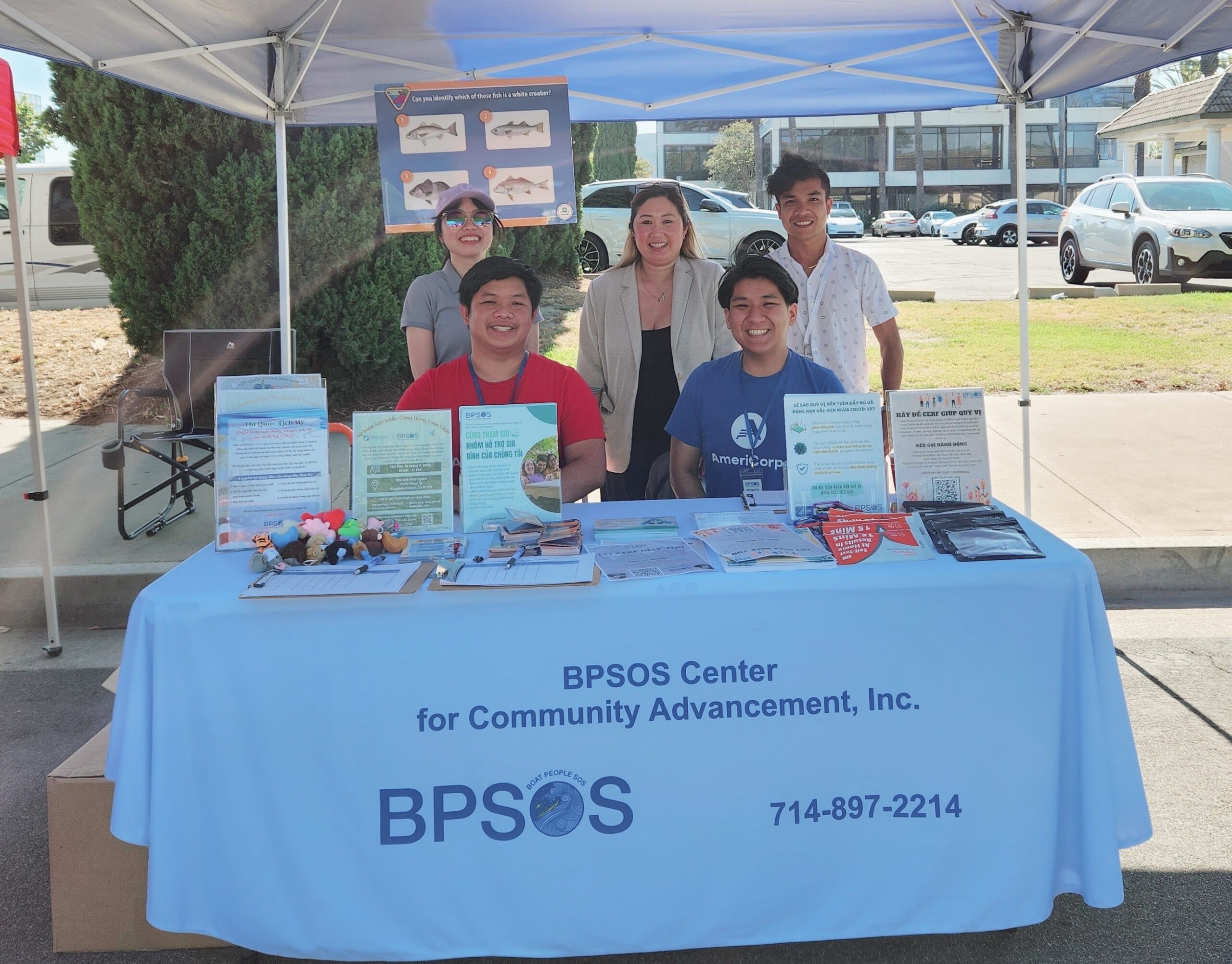 Five smiling adults, at a BPSOS table, are distributing information about vaccines and COVID-19 at an outdoor health care event.