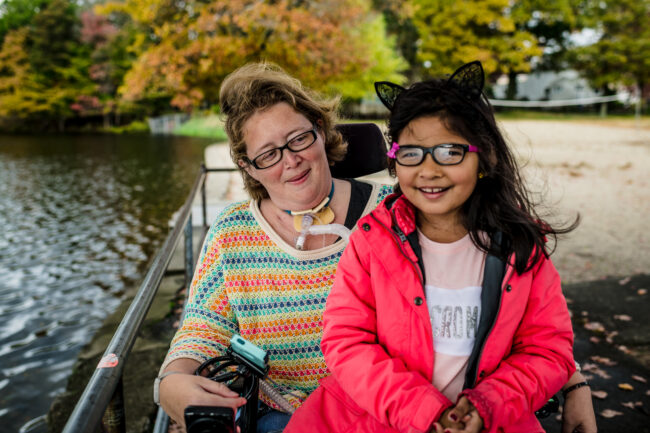 A mother wearing a multicolored shirt sits in a motorized wheelchair with a medical tube on her throat. She is posed with her child who is wearing a red raincoat and a pink and white shirt.