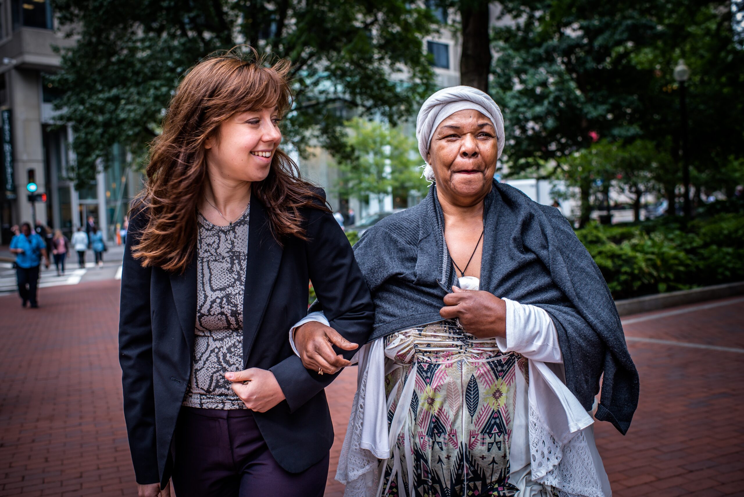 An older person and younger person walk through a metropolitan. The person on the left is dressed in a sheer white tunic, matching head wrap, and a beaded multicolored top with a stone necklace and sunglasses. The person on the right has brown hair and is wearing a necklace, black blazer and grey, and black top.