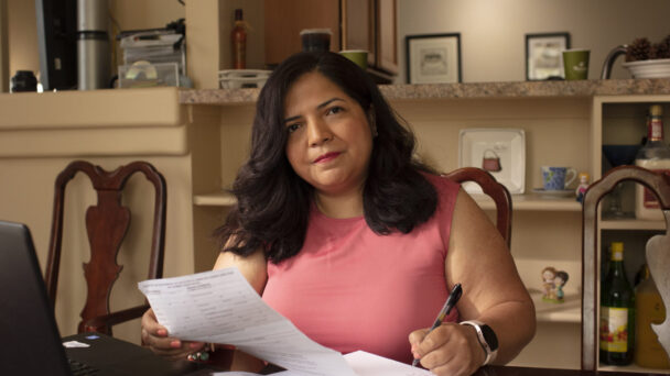 A woman with brown hair fills out forms related to her medical care. She is wearing a pink top and holding a pen while filling out forms next to her laptop. She is seated in the center of the photo at her kitchen table.