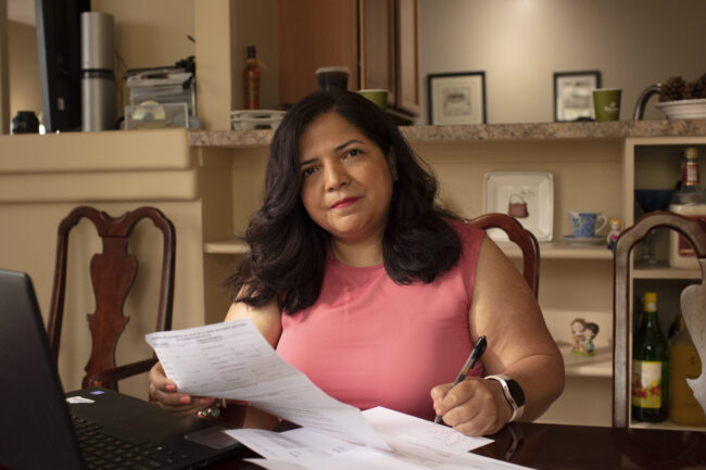 A woman with brown hair fills out forms related to her medical care. She is wearing a pink top and holding a pen while filling out forms next to her laptop. She is seated in the center of the photo at her kitchen table.
