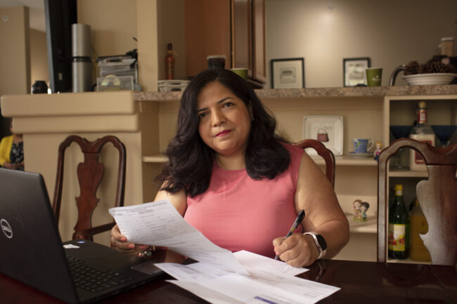 A woman with brown hair fills out forms related to her medical care. She is wearing a pink top and holding a pen while filling out forms next to her laptop. She is seated in the center of the photo at her kitchen table.