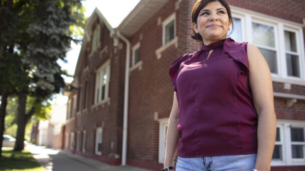 A person stands in front of a brick-covered apartment building. The person is smiling with a gaze to the right, and is wearing a burgundy top with blue jeans.