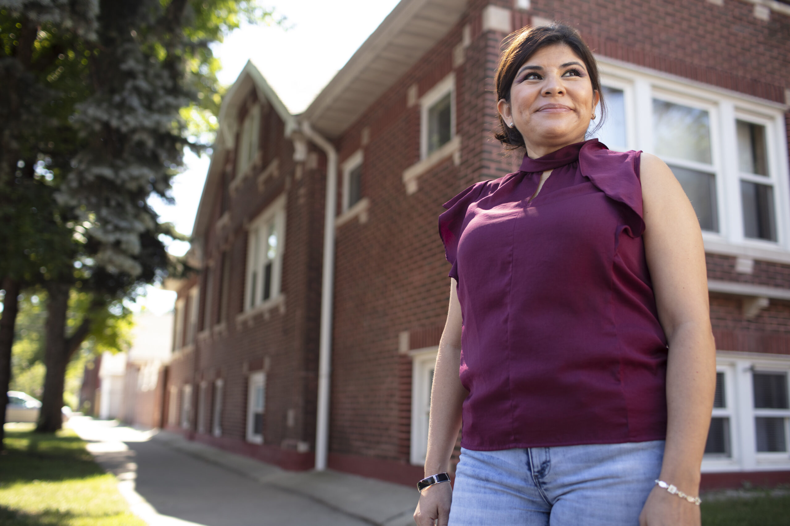 A person stands in front of a brick-covered apartment building. The person is smiling with a gaze to the right, and is wearing a burgundy top with blue jeans.