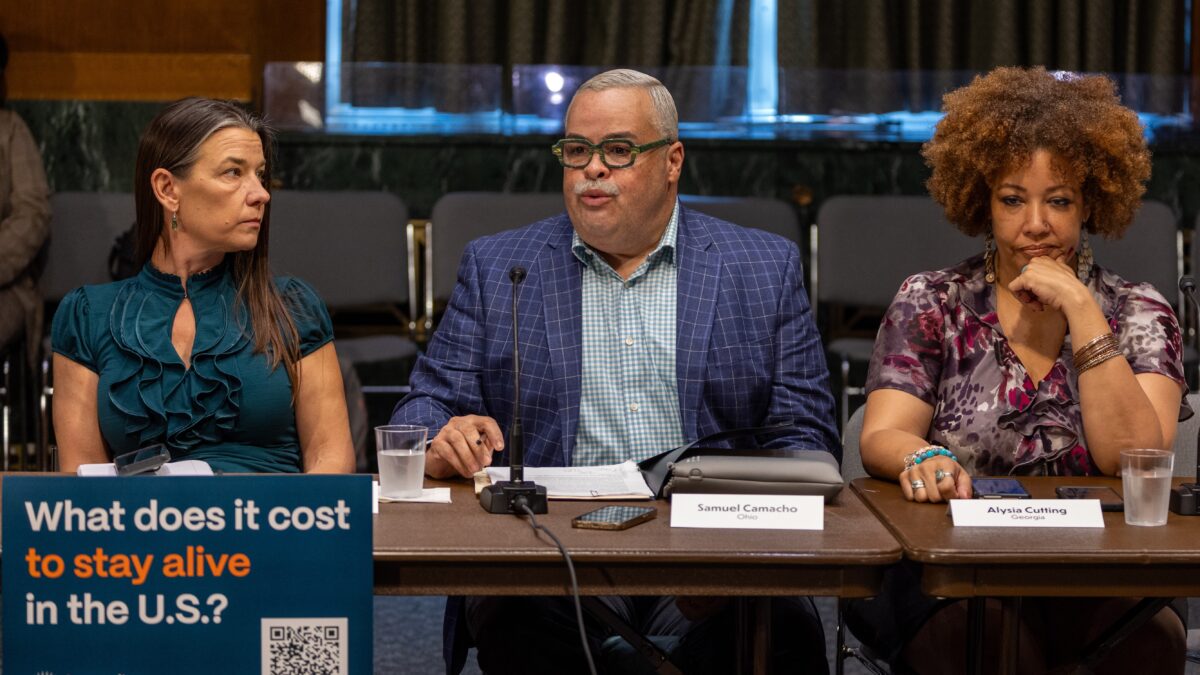 Three people sit at a table behind a sign that reads "What does it cost to stay alive in the U.S.?" The first speaker on the left has long dark hair and is wearing a blue top. The speaker in the middle has grey hair and is wearing glasses as well as a blue plaid top with a checkered shirt underneath. The last speaker on the right has dark hair and is wearing a purple and pink-toned multicolored shirt.