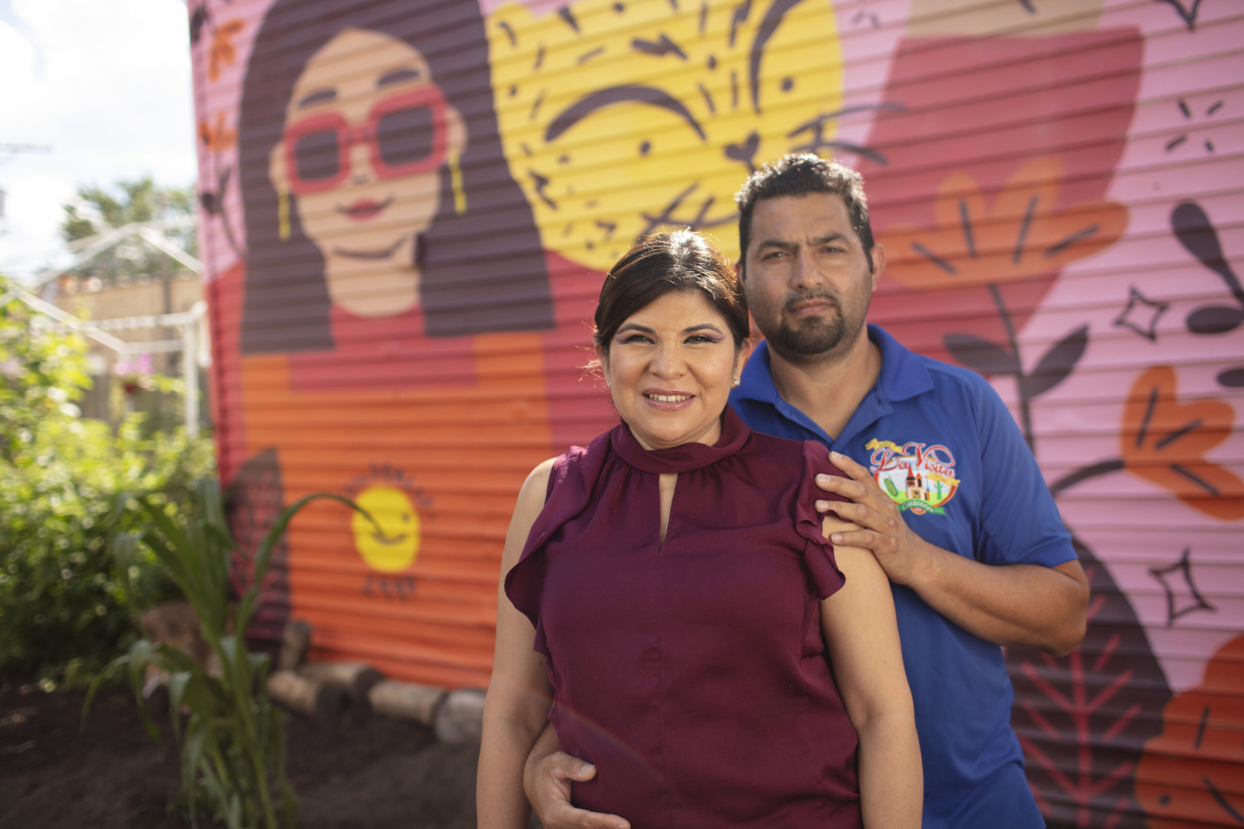 Sahida and her husband, Arturo, pose in front of a brightly colored mural and garden. Sahida is posed in front with a burgundy, sleeveless top, while her husband, standing behind her, embraces her while wearing a blue top with a small multicolored logo.