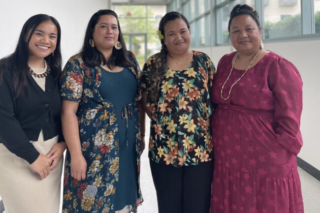 A group of four people pose in an office hallway. From left to right: person is wearing a black top and beige dress with a necklace. The second person is wearing earrings, a long blue tunic top with black pants and a multi-colored robe. The third person is wearing a top with an assortment of flower graphics and black pants and a flower hair clip. The fourth person is wearing a pink dress with a necklace.