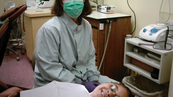 A dentist poses in a dentist office with a young patient receiving dental care. The dentist in the background is wearing a mask, gloves, and a dentist uniform. The young patient is lying down on a dentist chair wearing glasses and is smiling with a napkin covering her shirt.