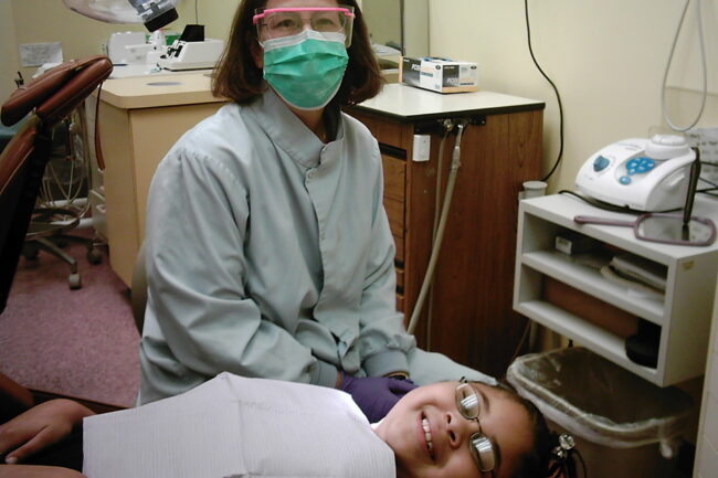 A dentist poses in a dentist office with a young patient receiving dental care. The dentist in the background is wearing a mask, gloves, and a dentist uniform. The young patient is lying down on a dentist chair wearing glasses and is smiling with a napkin covering her shirt.