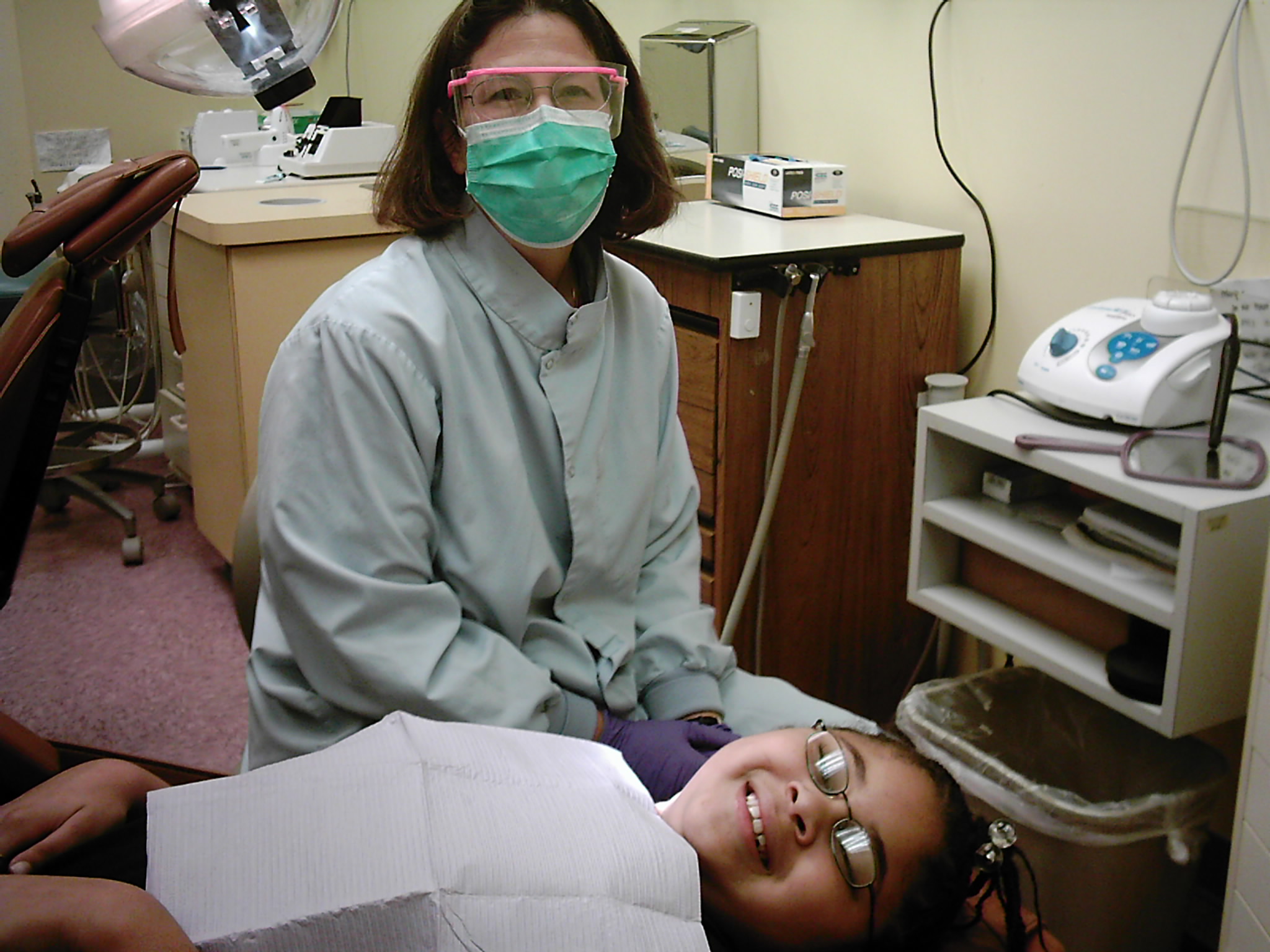 A dentist poses in a dentist office with a young patient receiving dental care. The dentist in the background is wearing a mask, gloves, and a dentist uniform. The young patient is lying down on a dentist chair wearing glasses and is smiling with a napkin covering her shirt.