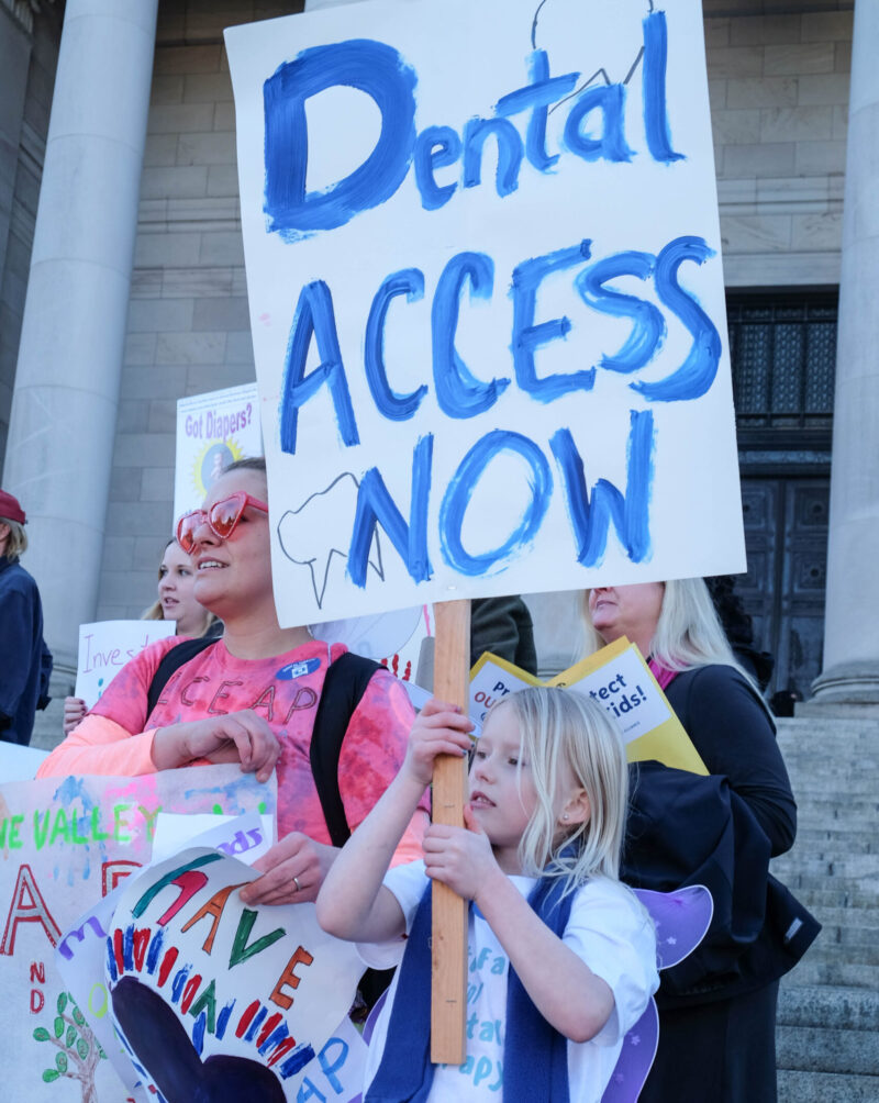A child with shoulder length blonde hair holds a large sign saying "Dental Access Now" in front of a couple other people.