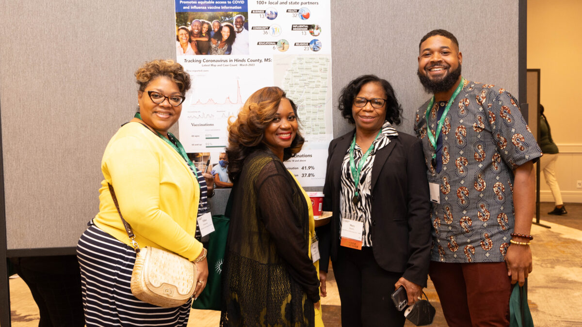 A group of four people pose for a photo in front of an informational poster in a conference room.